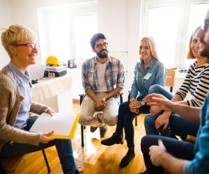 Group of people sitting in a circle on group therapy. Looking at their therapist and listening ti her story.